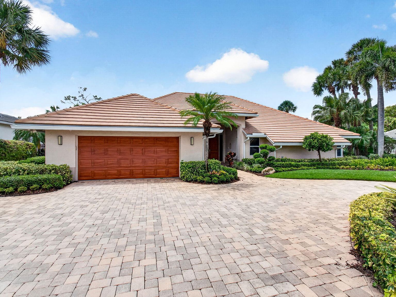 The paver driveway curving up to the two-car garage, anchored by a single palm in the courtyard planter
